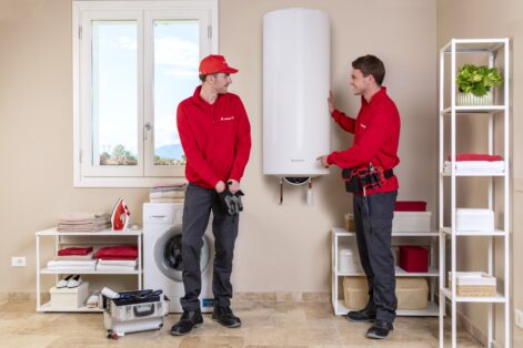 Due tecnici Ariston installano uno scaldabagno verticale in una lavanderia ordinata, con scaffali e lavatrice